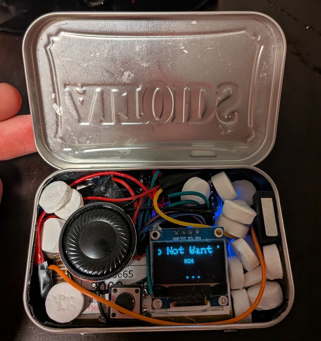 An open Altoids tin holding Cam Smith's mintp3 cyberdeck — a speaker, small OLED screen showing a song title, and messy wiring nestled among mints.