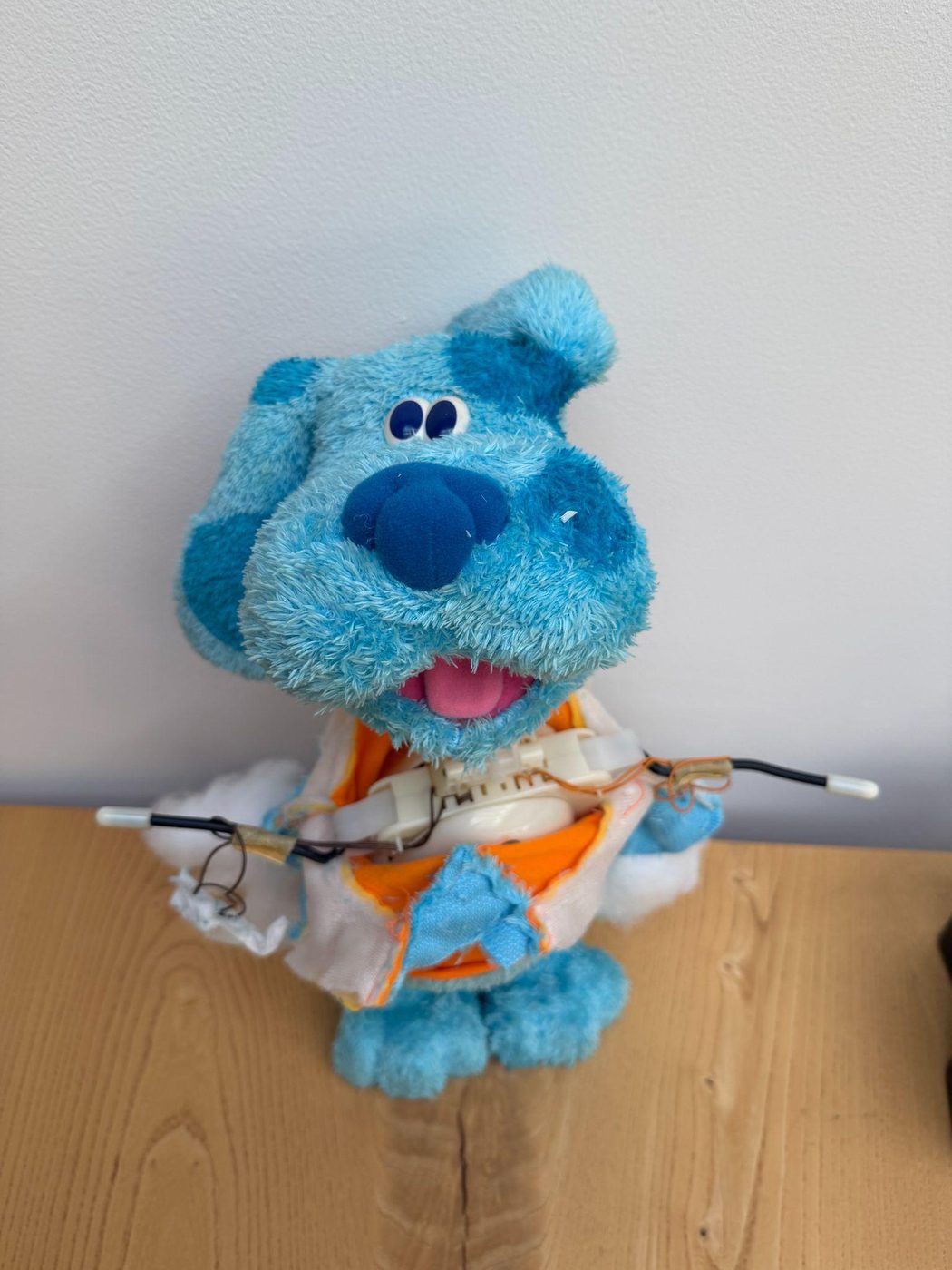 A blue plush toy dog with its chest carefully opened, small wires and mechanism visible inside, sitting on a wooden table.