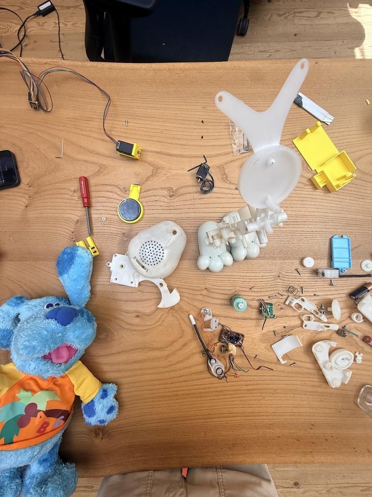 Overhead view of a plush toy on the left and everything that was inside it laid out on a wooden table — motors, speaker, small chassis pieces, wires, buttons.