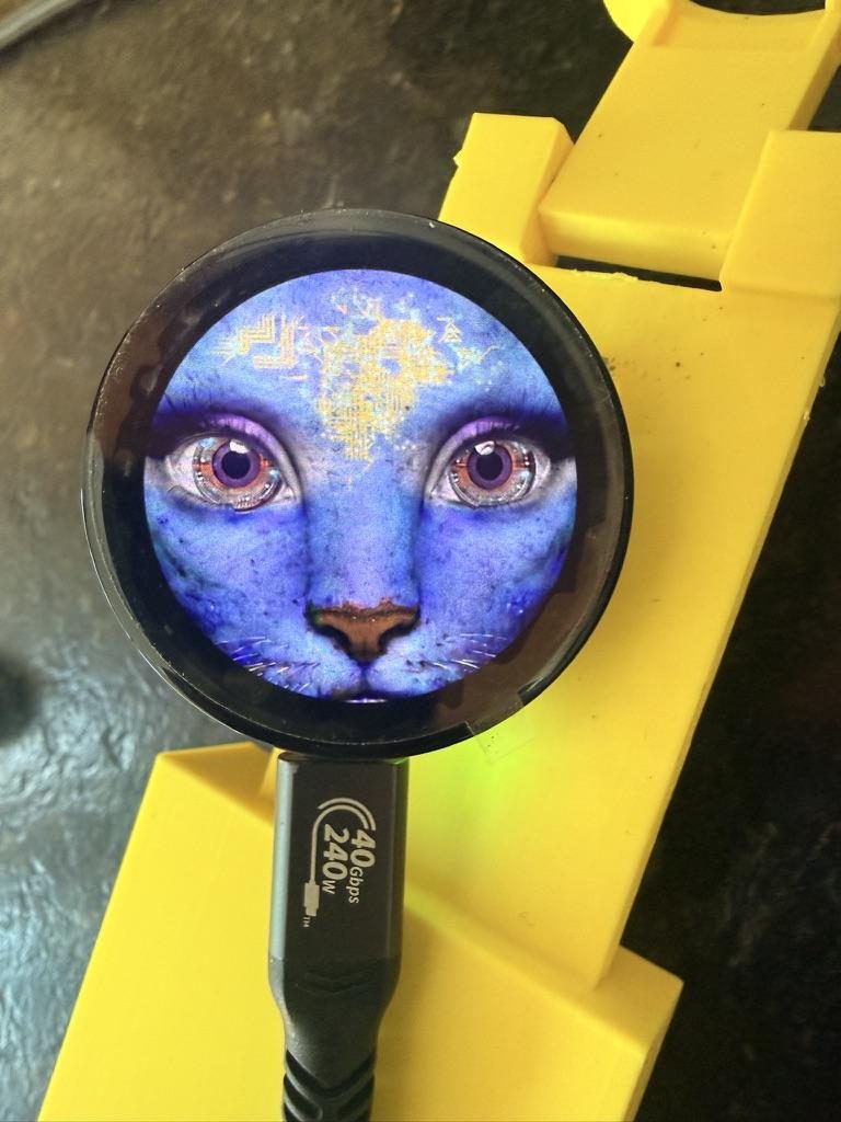 Close-up of a small round screen fitted into a 3D-printed yellow mount, displaying an illustrated blue cat face with vivid painted eyes.