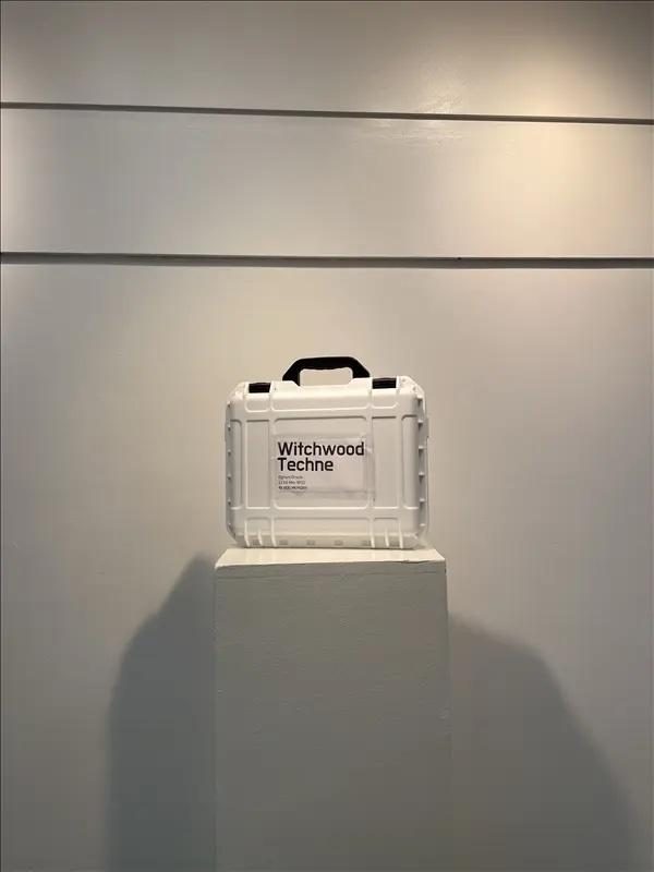 A closed white Pelican case labelled Witchwood Techne, on a plinth in a gallery.