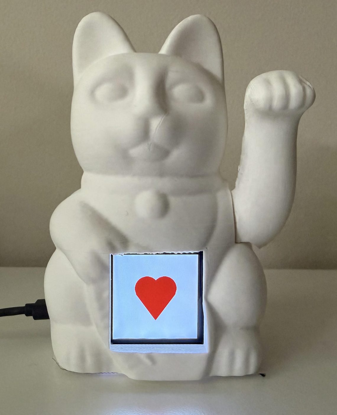 A white ceramic waving-cat figurine with a small screen in its belly, displaying a red heart. Its paw is raised.