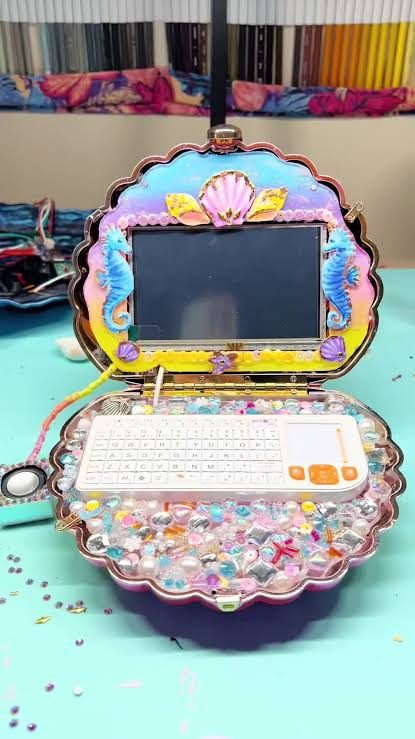 A pink seashell-shaped handheld computer, encrusted with beads and rhinestones, with a small screen showing seahorses.