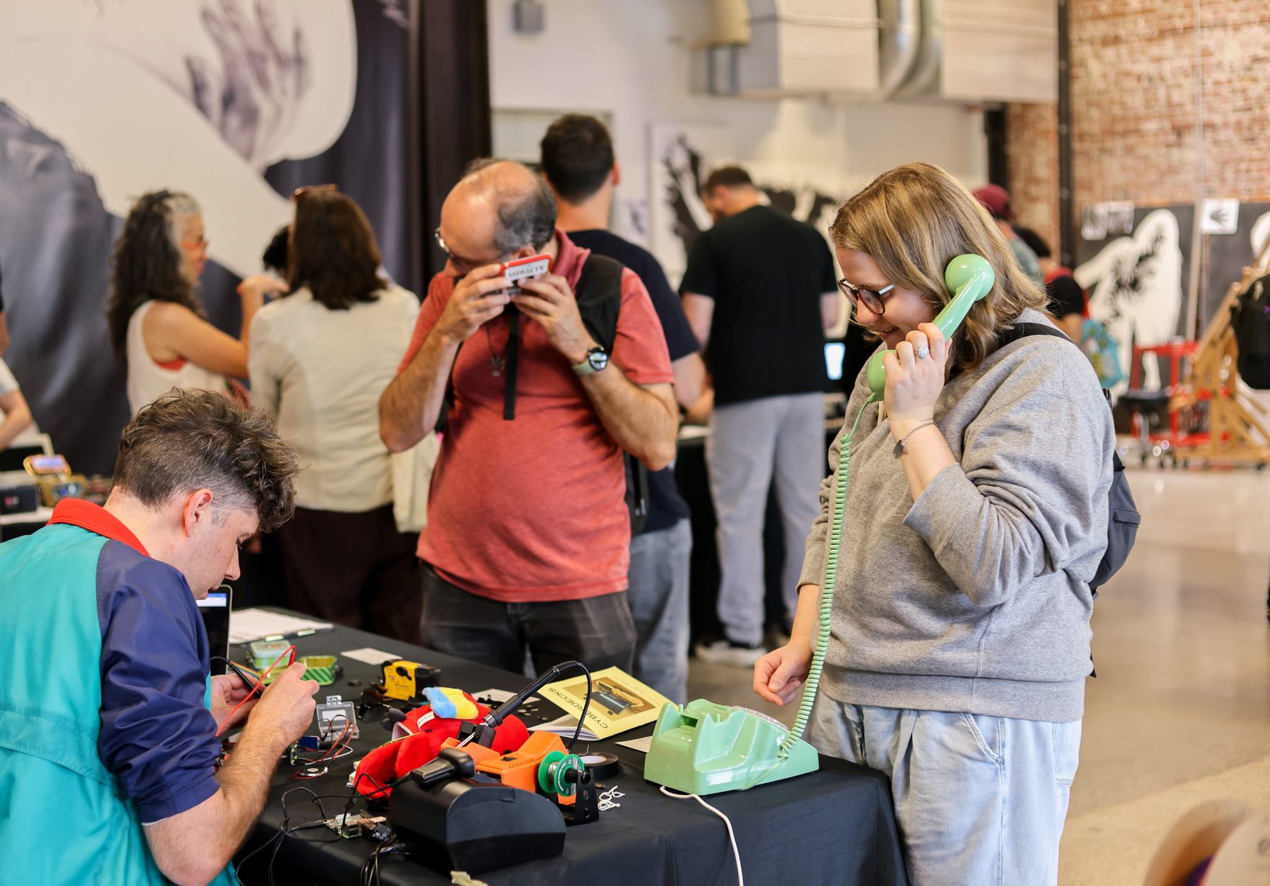 A visitor listens on a green rotary phone at a table of cyberdecks; another visitor mid-solder in the foreground.