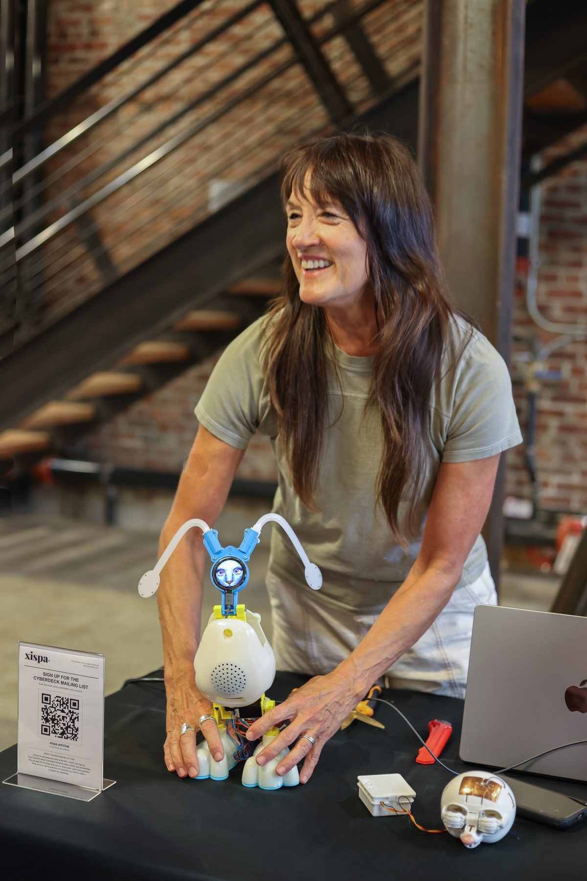 Cathy McClure with ChimeraBot, a 3D-printed white creature-cyberdeck, at her table.