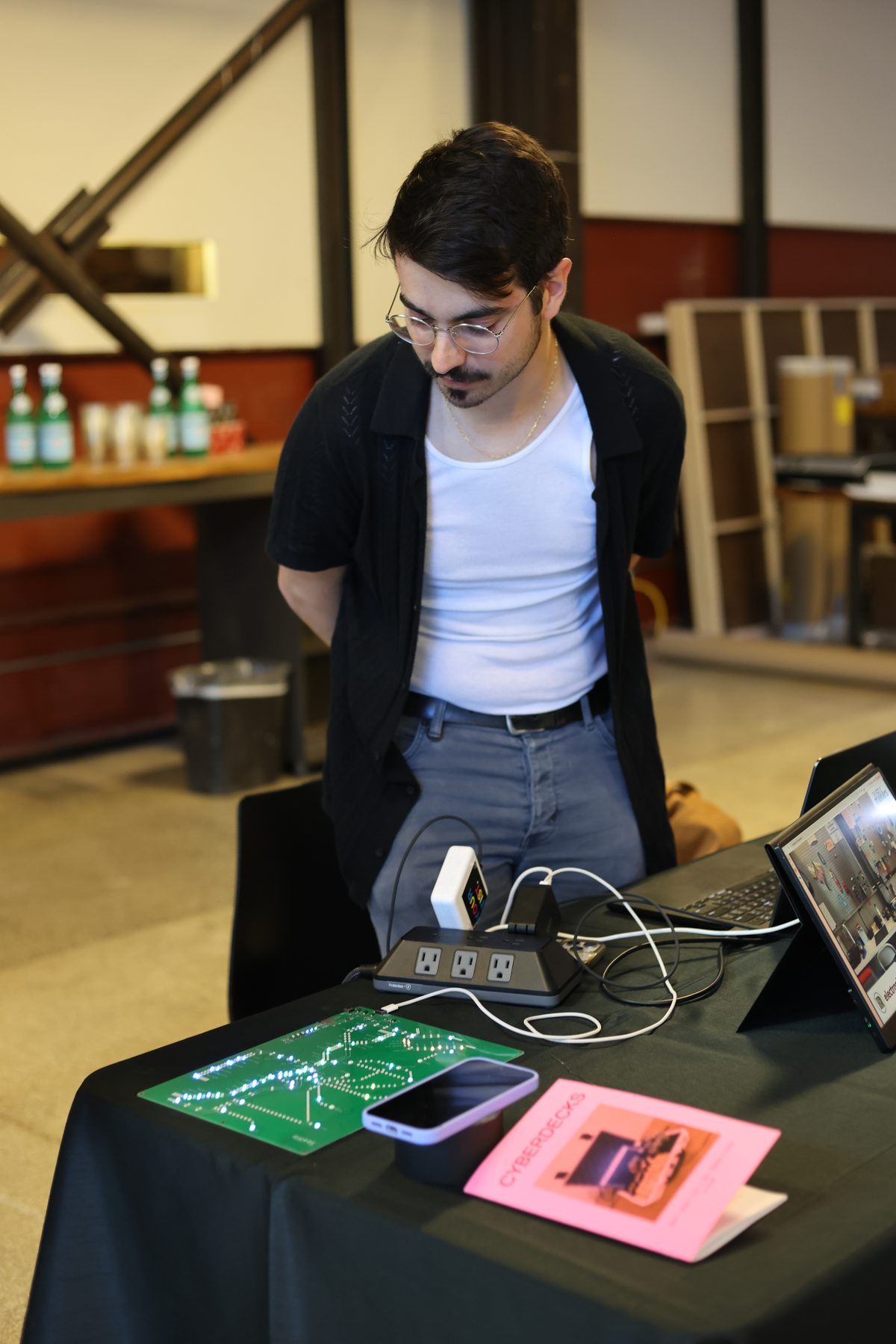 Cam Smith at his table, a green PCB cyberdeck and copies of the Cyberdecks zine spread in front.