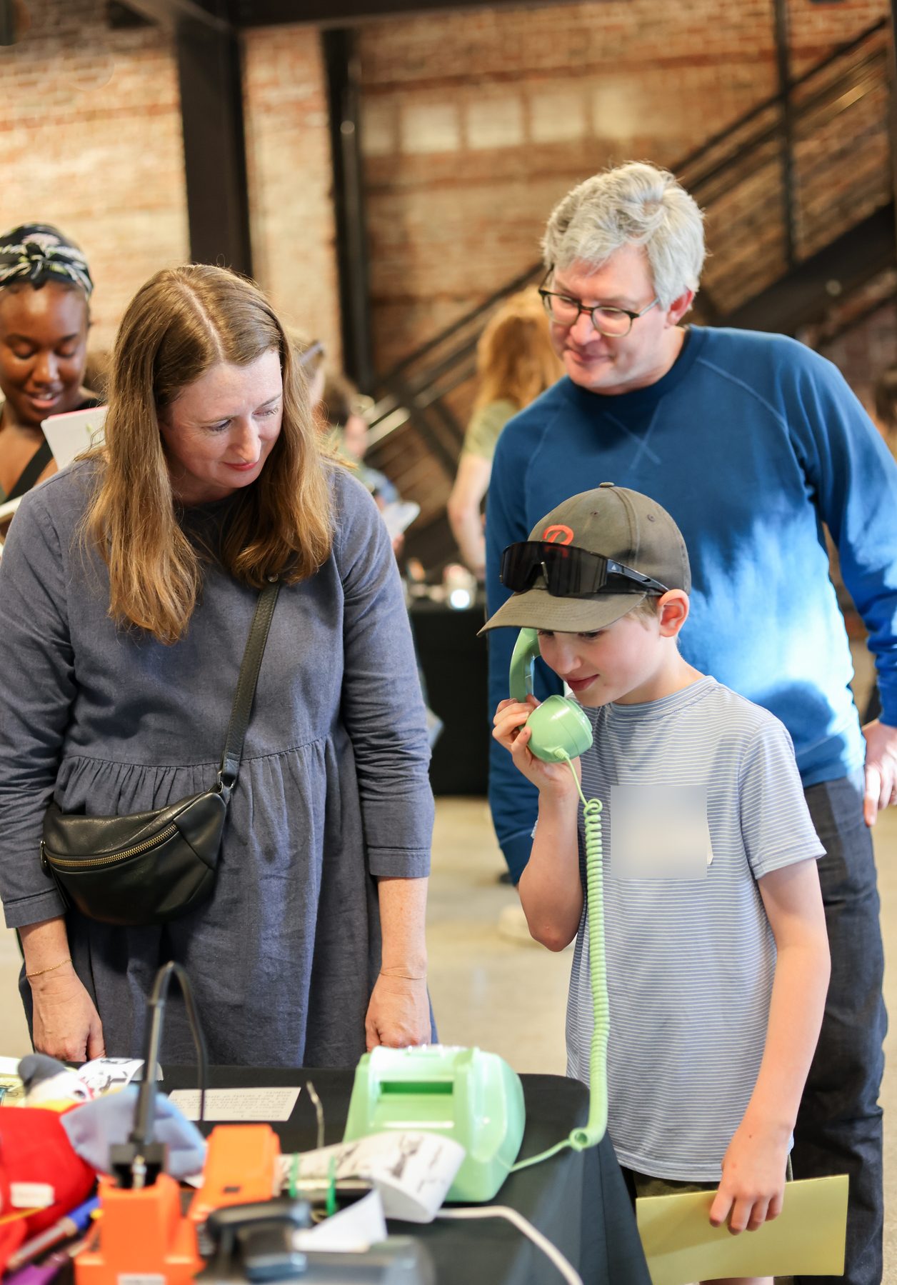 A young visitor with a green telephone handset, parents watching, at a table of decks.