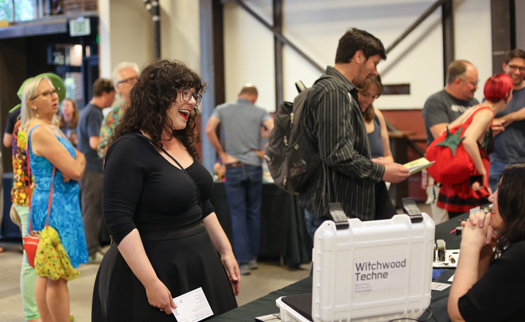 Kate Bailey laughing beside the Witchwood Techne Pelican-case cyberdeck; the room full of people behind her.